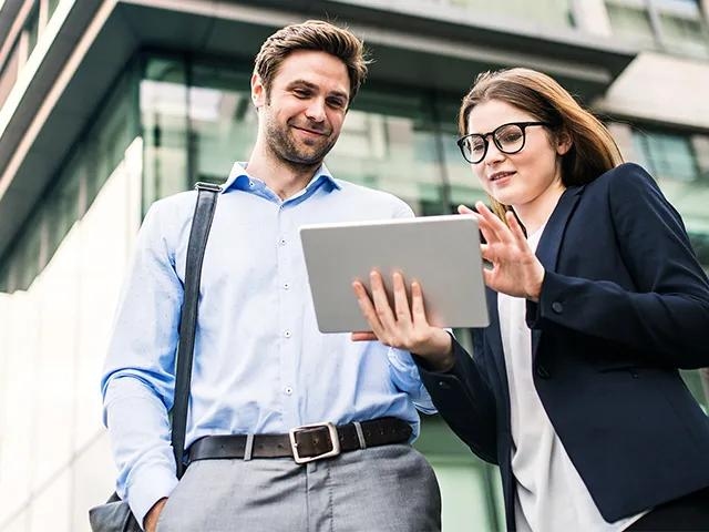 Zwei Personen stehen vor einem modernen Bürogebäude und betrachten gemeinsam ein Tablet.